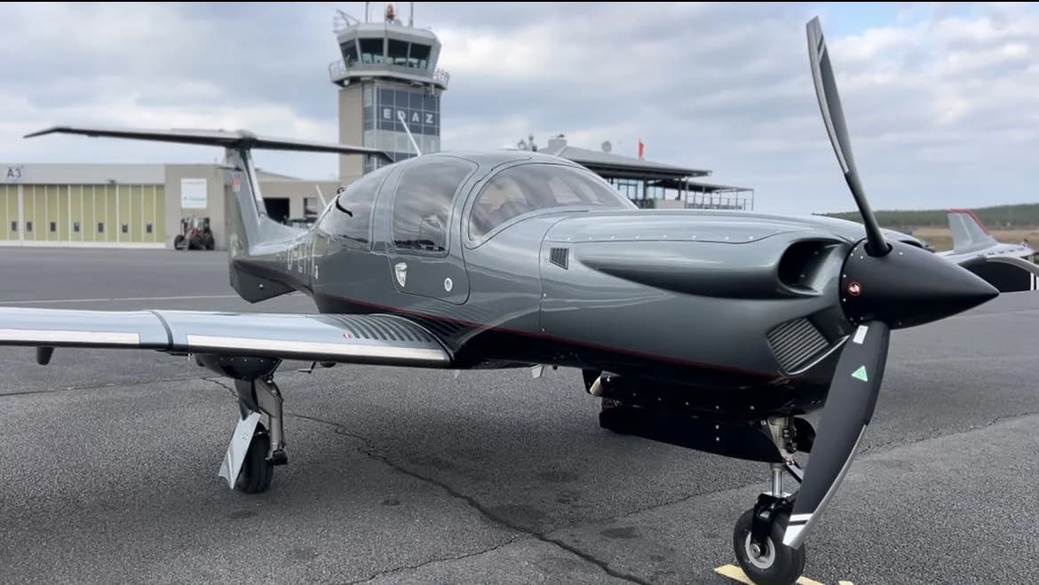 Close-up of a gray Diamond DA50 RG aircraft parked on the tarmac with an airport control tower in the background.