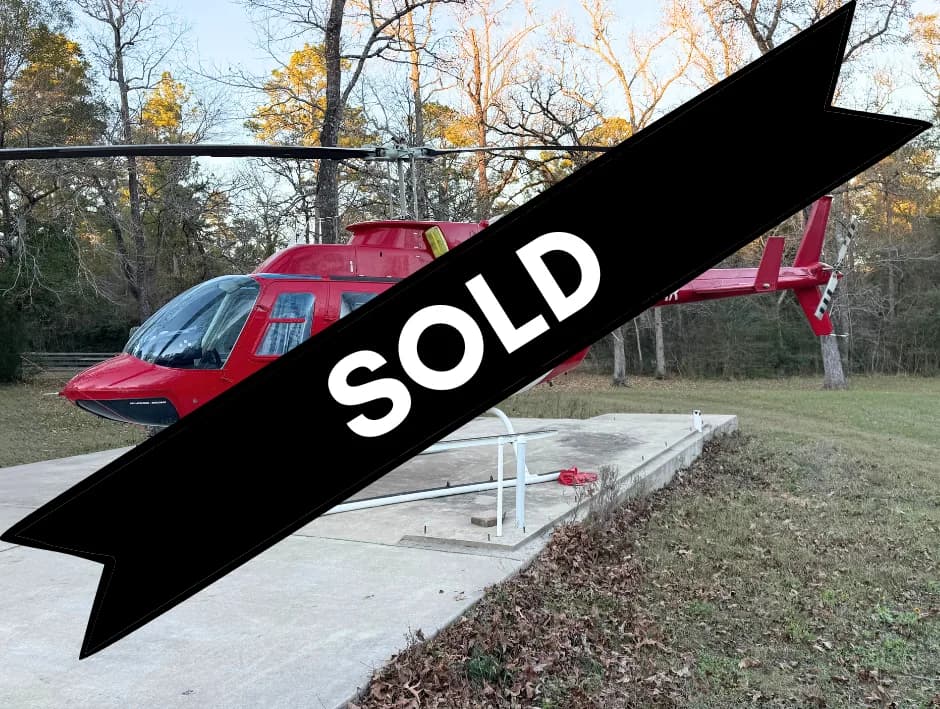A red helicopter with a clear bubble canopy parked on a concrete pad in a wooded area.