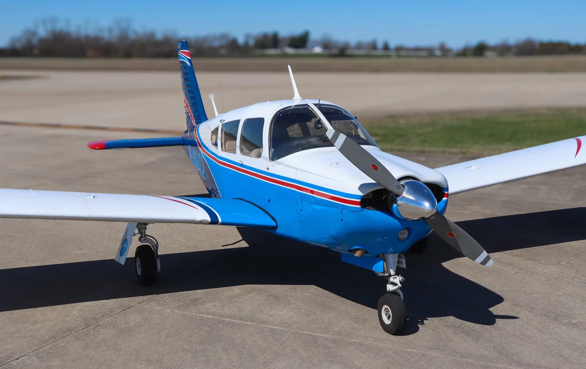 A front-quarter view of a blue and white Piper Cherokee aircraft with red stripes parked on an airfield.
