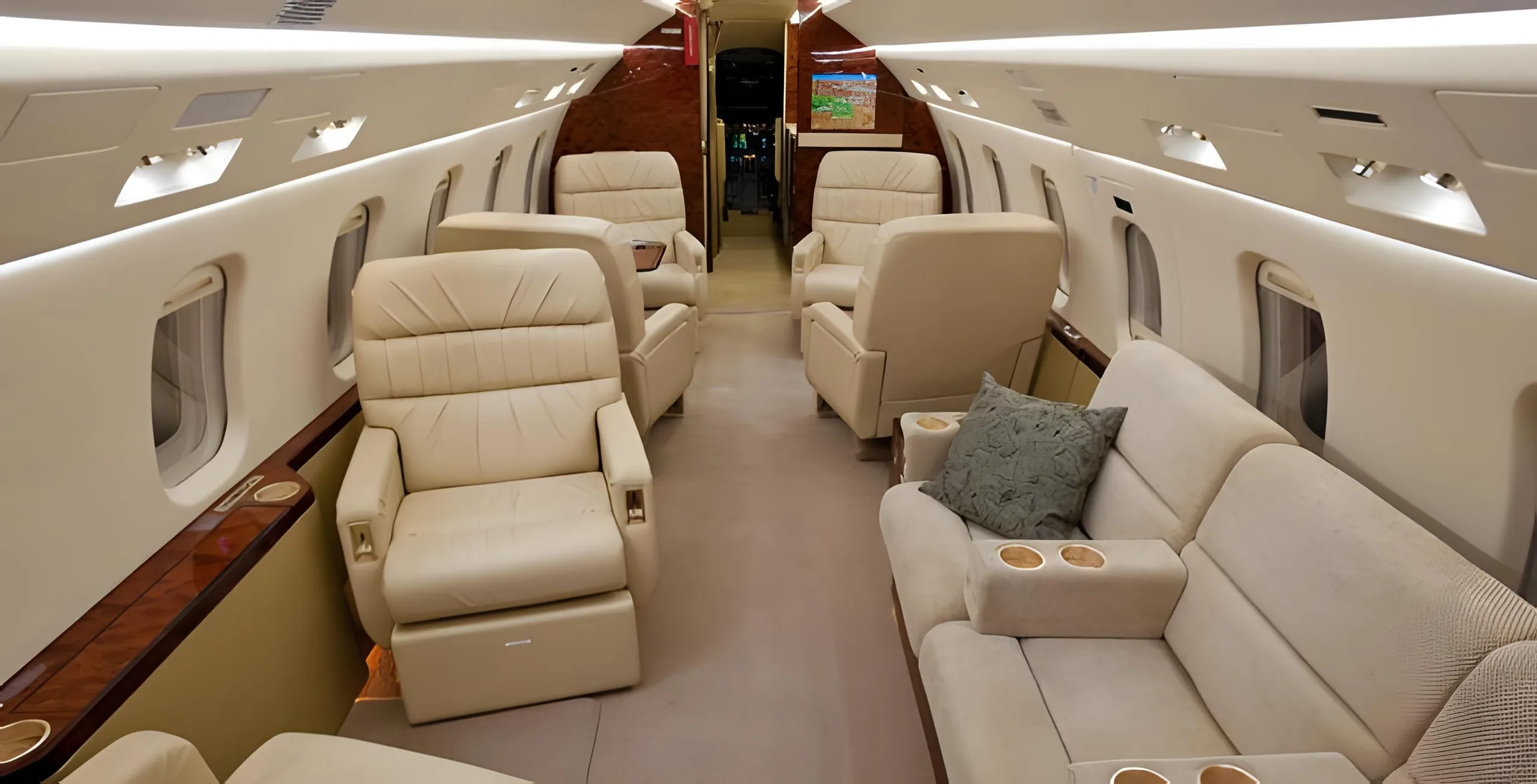 A wide view of the aircraft's spacious cabin interior featuring multiple beige leather seats and a light-colored sofa, with a map displayed on a screen at the rear.