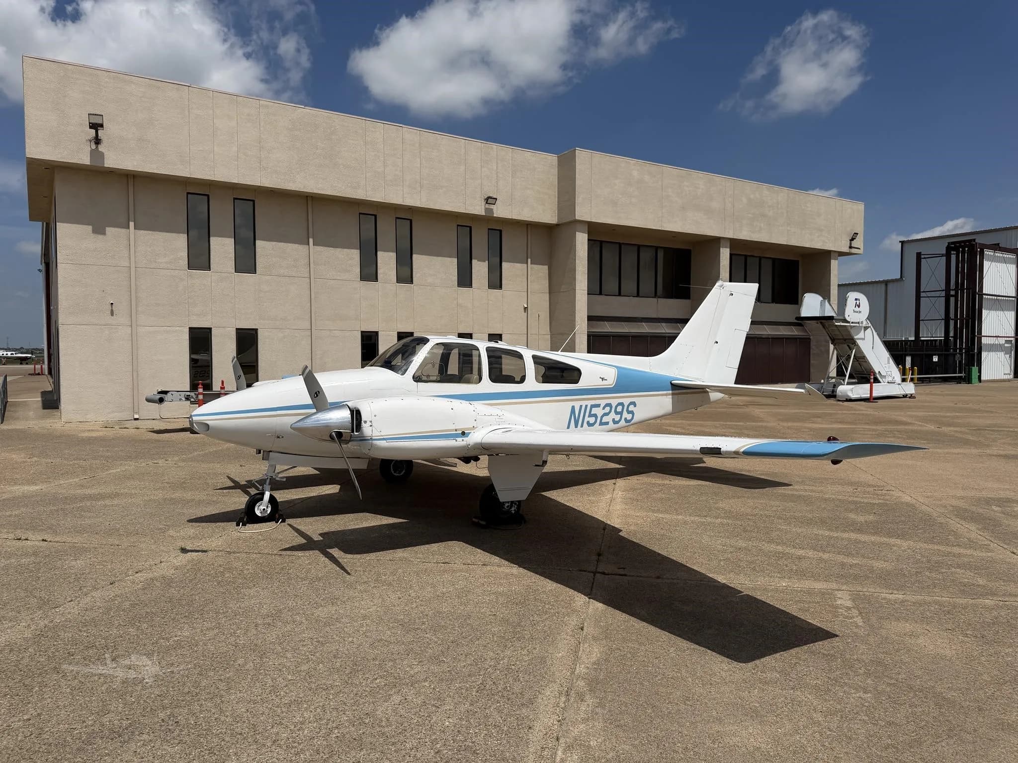 A white and blue twin-engine propeller aircraft, N1529S, parked on an apron in front of a beige building under a partly cloudy sky.