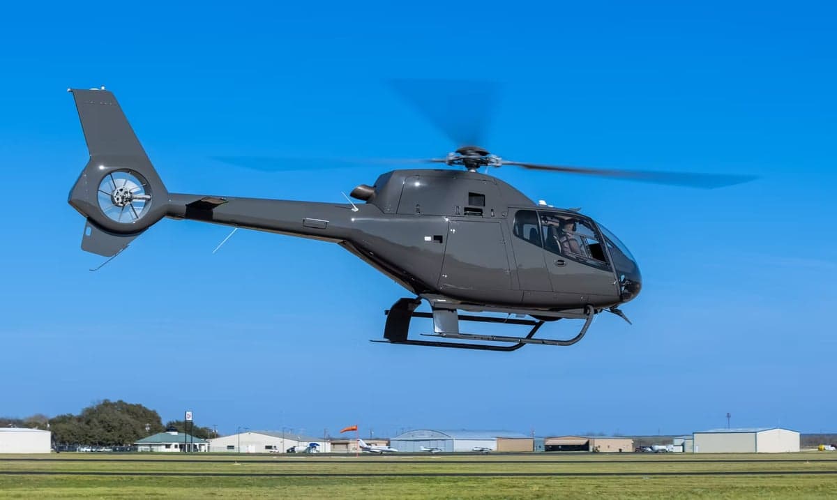 A gray helicopter taking off from a grassy airfield.