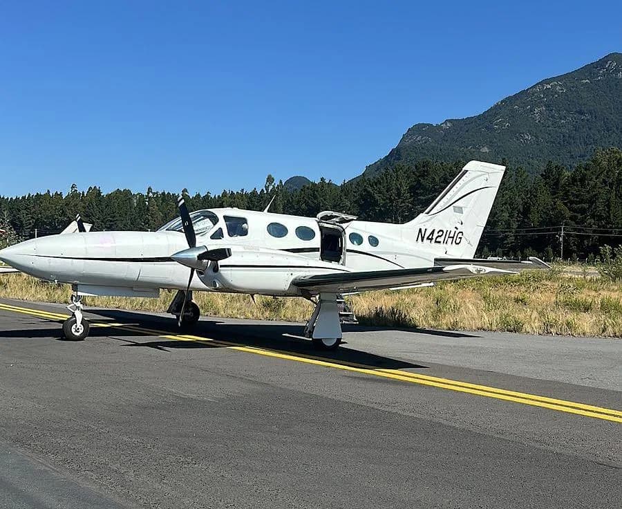 A white Cessna 421 Golden Eagle twin-engine aircraft with registration N421HG parked on an airfield with mountains in the background.