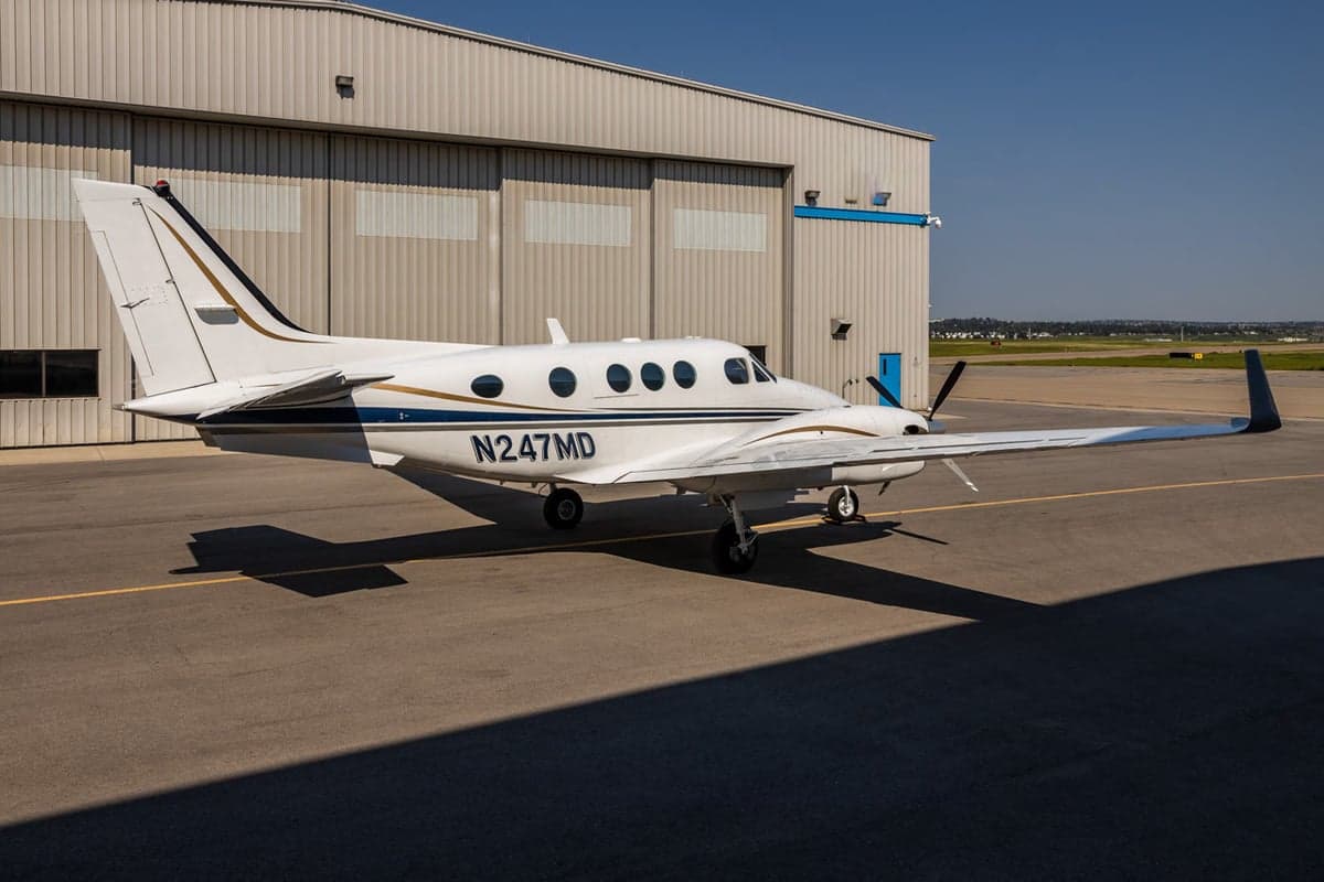 Side view of a white and blue Beechcraft King Air 90 with registration N247MD parked in front of a hangar.