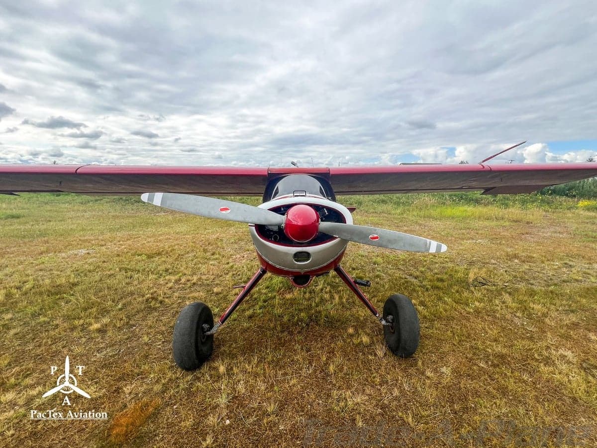 A head-on view of a red and silver single-engine aircraft with a propeller and large tires, parked on a grassy field.