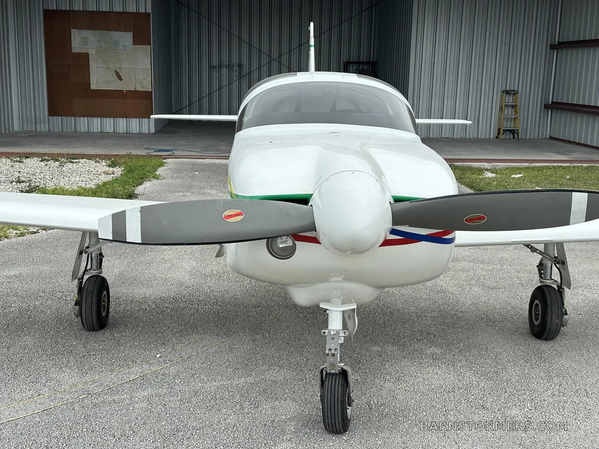 Front view of a white single-engine propeller aircraft with green and red/blue stripes, parked in front of a hangar.