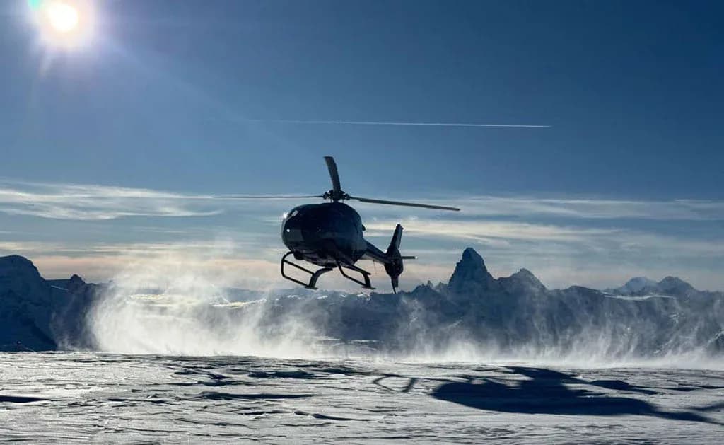 A dark helicopter hovers low over a snowy, windswept mountain landscape under a bright sun.