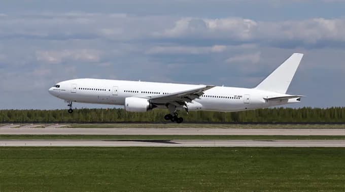 A large, all-white Boeing 777 aircraft with landing gear extended, flying low over a runway with green grass.