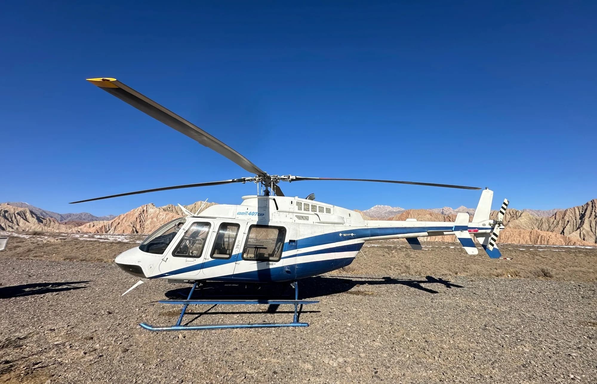 A white and blue Bell 407GXP helicopter is parked on a gravel field with mountains in the background under a clear blue sky.