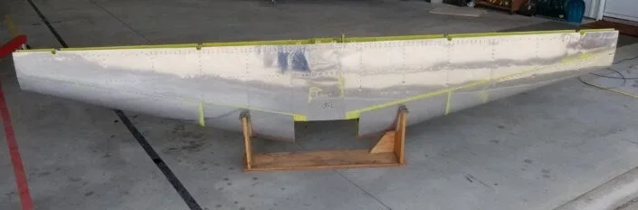 A partially assembled aircraft wing section, showing its metal skin, on wooden stands.