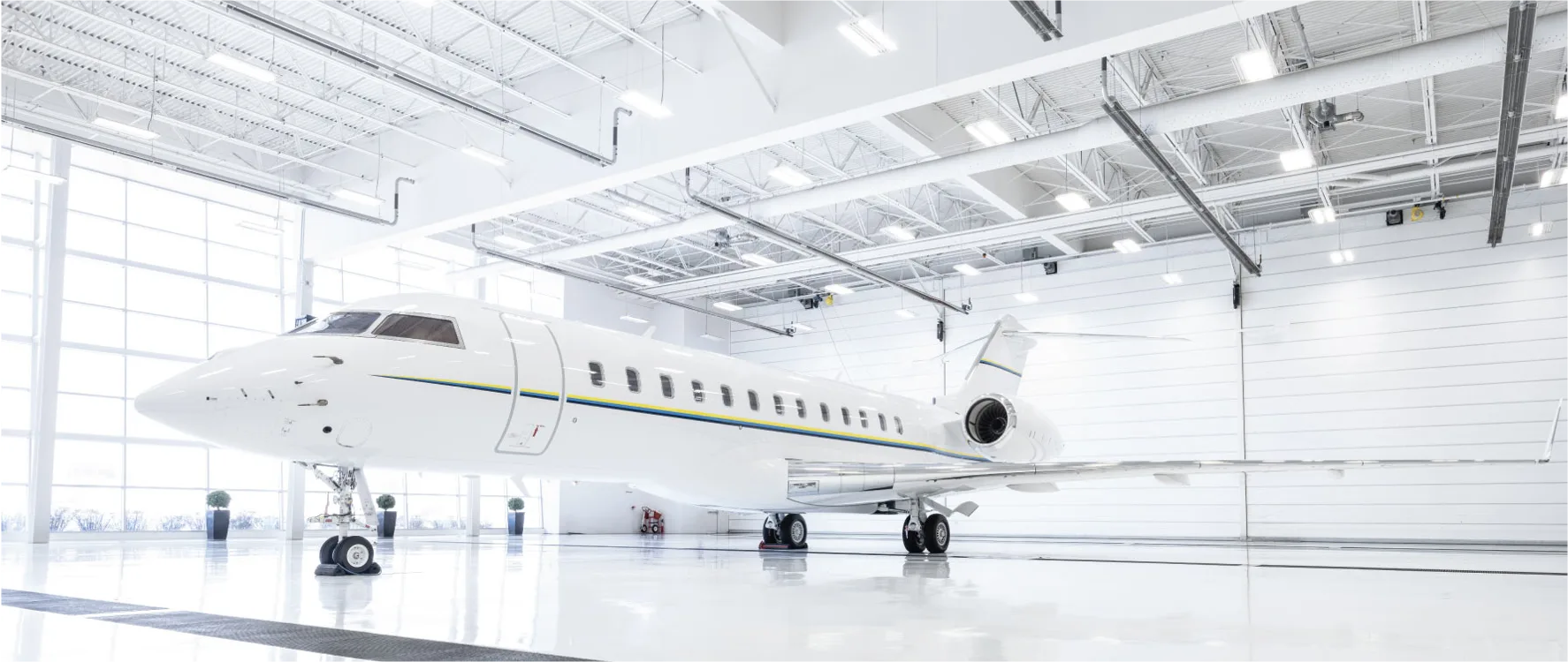 A white private jet with a blue and yellow stripe sits in a brightly lit, modern hangar.