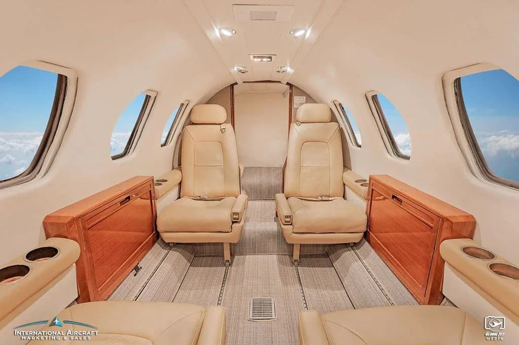 The mid-cabin interior of a business jet featuring beige leather seats, wooden side consoles, and windows showing clouds.