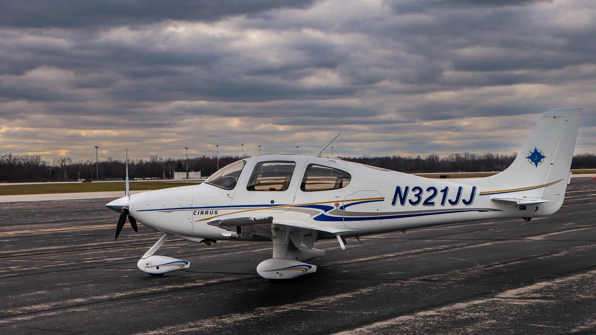 A white Cirrus aircraft, registration N321JJ, with blue and gold stripes, is parked on a tarmac under a cloudy sky.