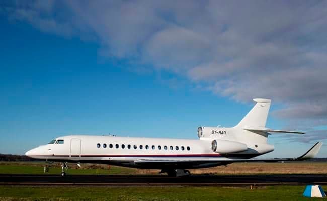 A white Dassault Falcon business jet with registration OY-RAD is parked on a tarmac under a bright blue sky.