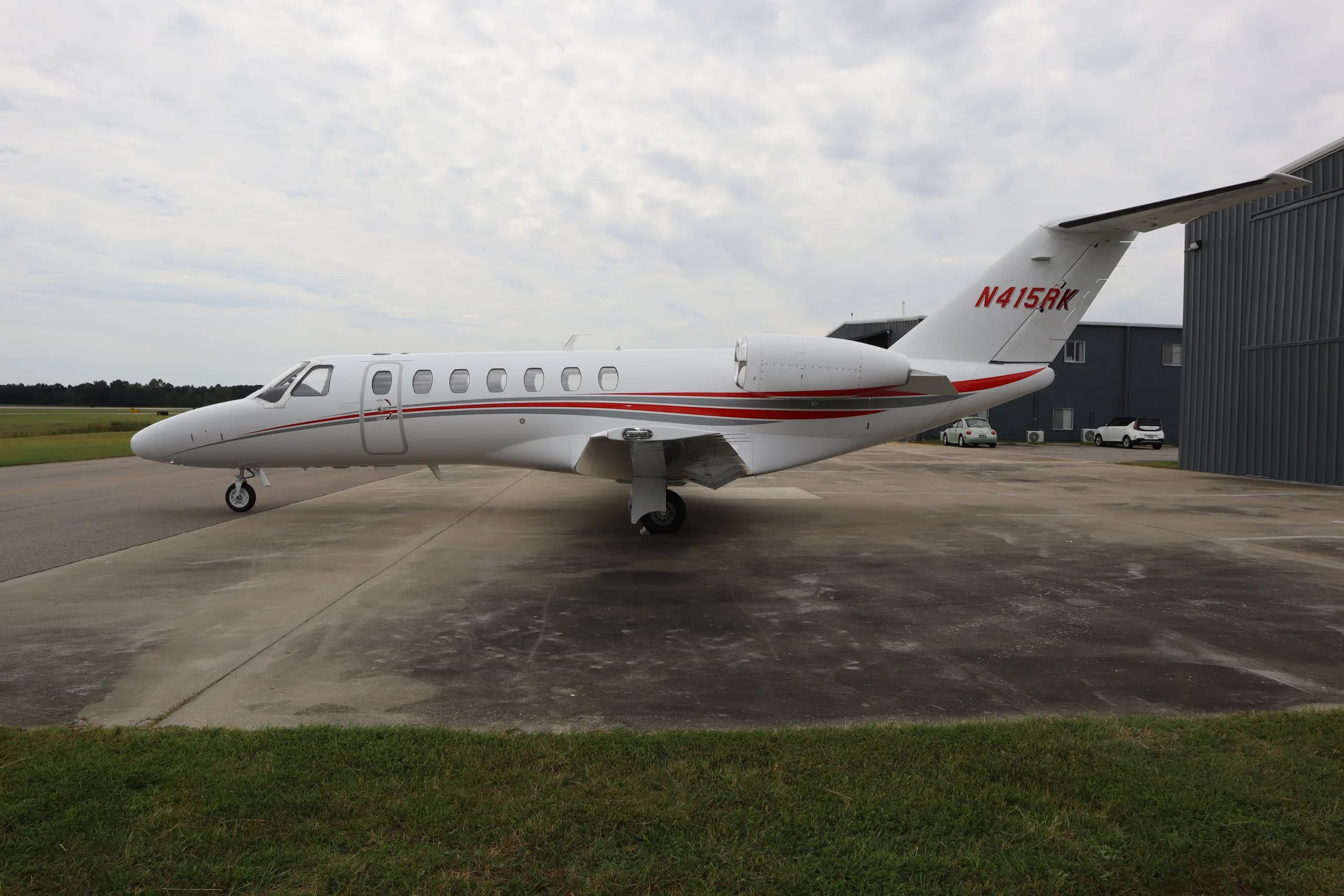 A side view of the white Cessna Citation business jet N415RK with red and grey stripes, parked on an tarmac with grass in the foreground.