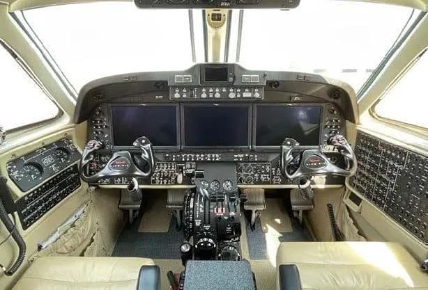 The cockpit of an aircraft featuring three large display screens, dual control yokes, and a central throttle quadrant.