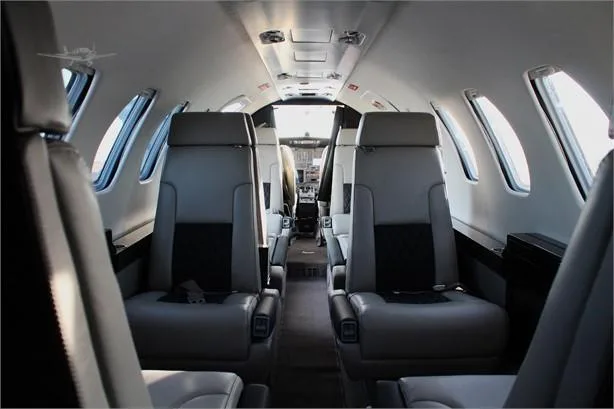 A view down the aisle of a private jet cabin, showing light gray and black seats leading to the cockpit.