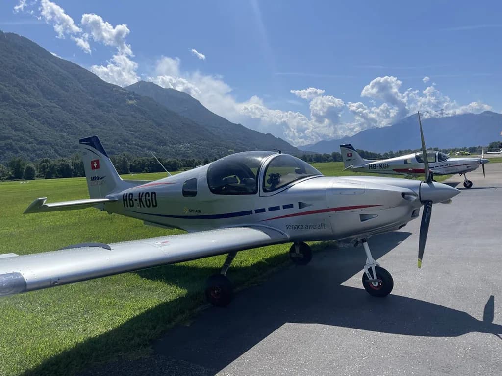 A silver Sonaca 200 aircraft (HB-KGD) with blue and red stripes is parked on an airfield with mountains in the background.