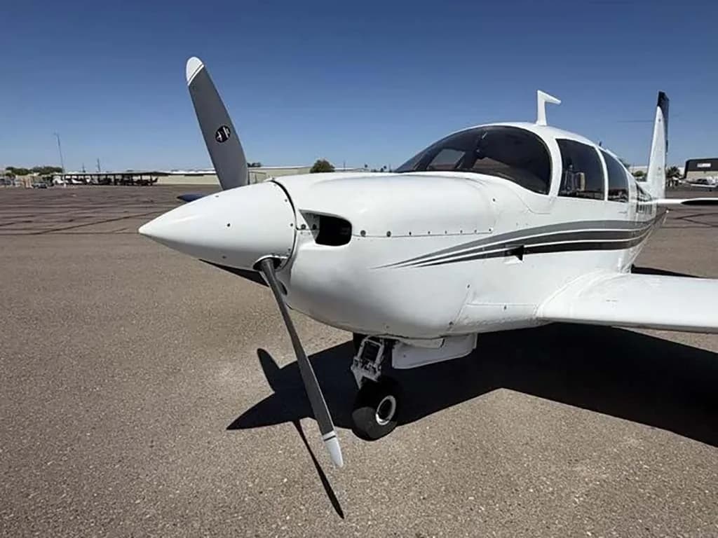 A close-up view of the front of a white Mooney M20 aircraft with a single propeller on an airfield.
