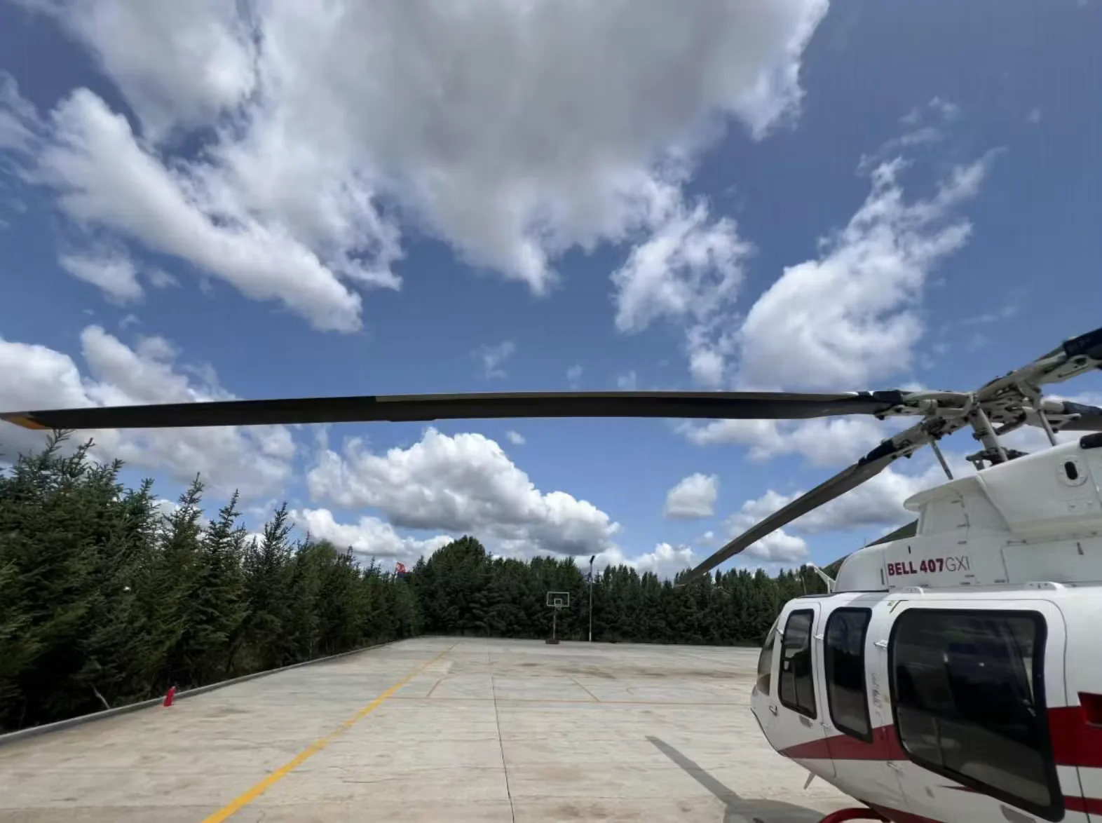 A close-up view of the main rotor and fuselage of a white and red Bell 407 GXi helicopter on a concrete pad.