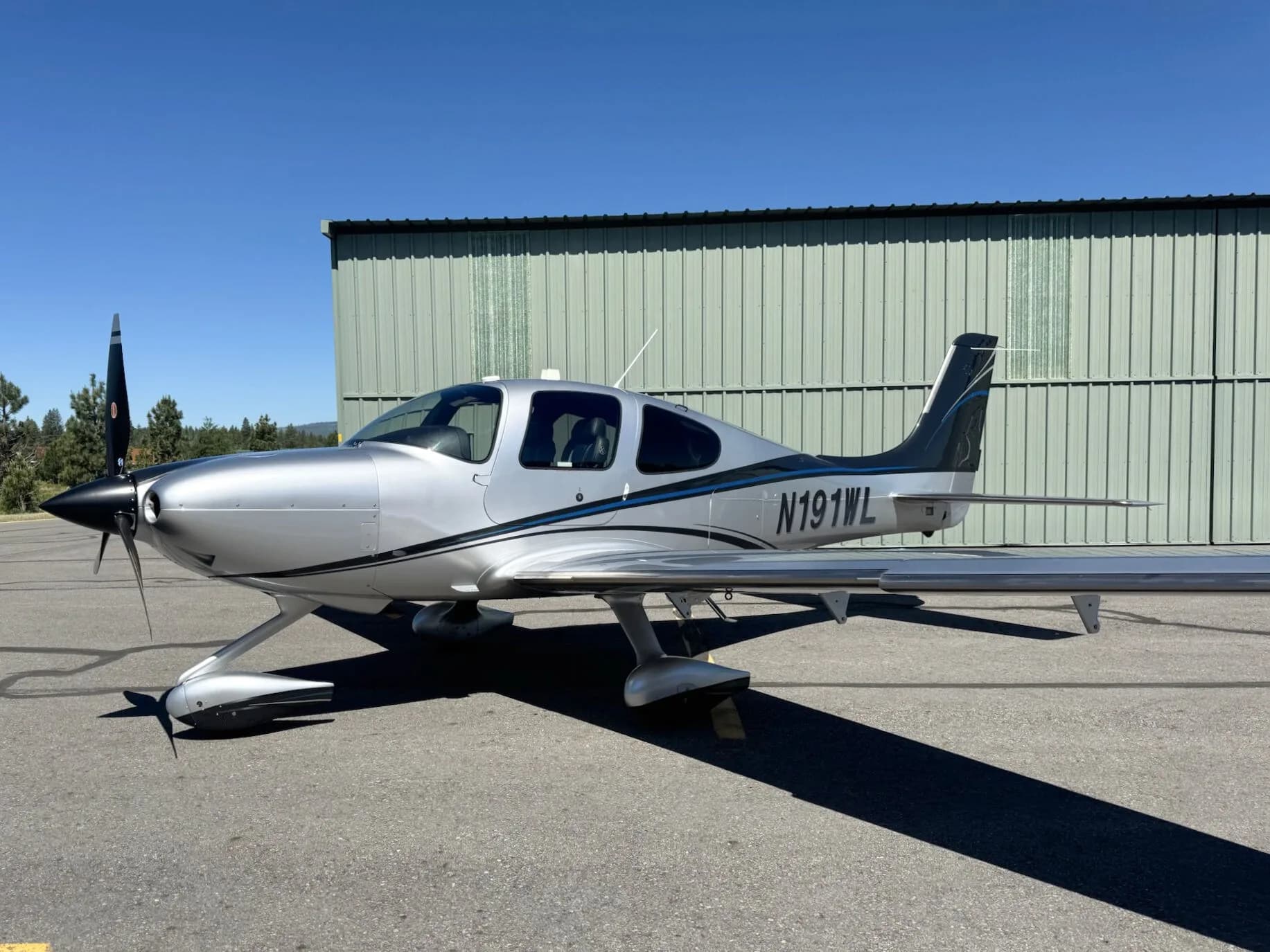 A side profile view of a silver and black Cirrus SR22T aircraft with N191WL, parked on an asphalt apron next to a hangar.