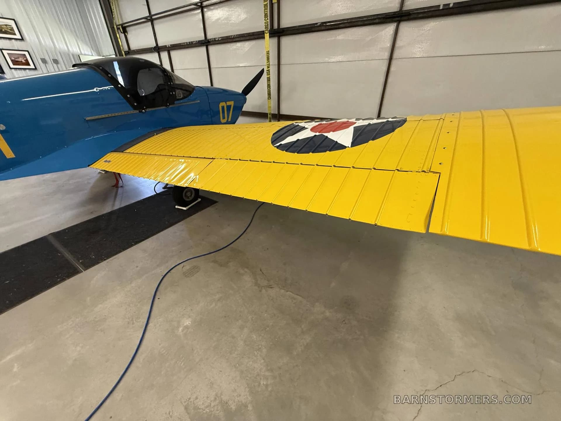 Close-up of the yellow wing with a US star insignia and the blue fuselage of a single-engine aircraft in a hangar.