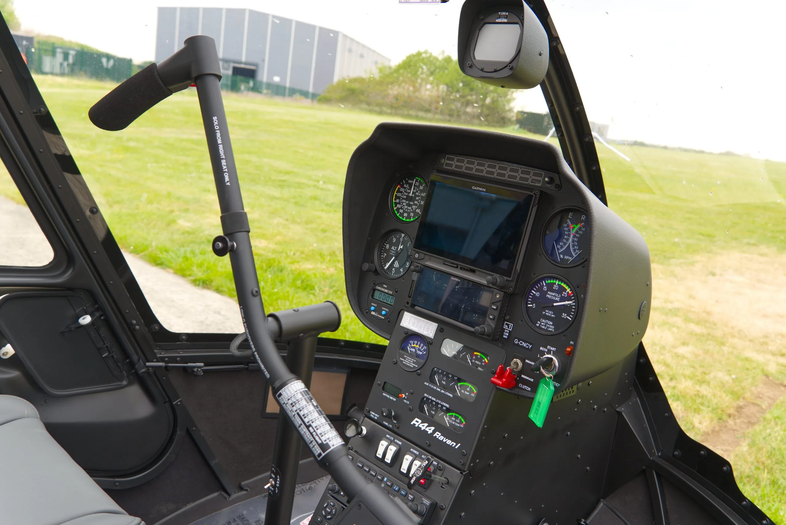 The black instrument panel and controls of a Robinson R44 Raven I helicopter, with a green field visible through the windshield.