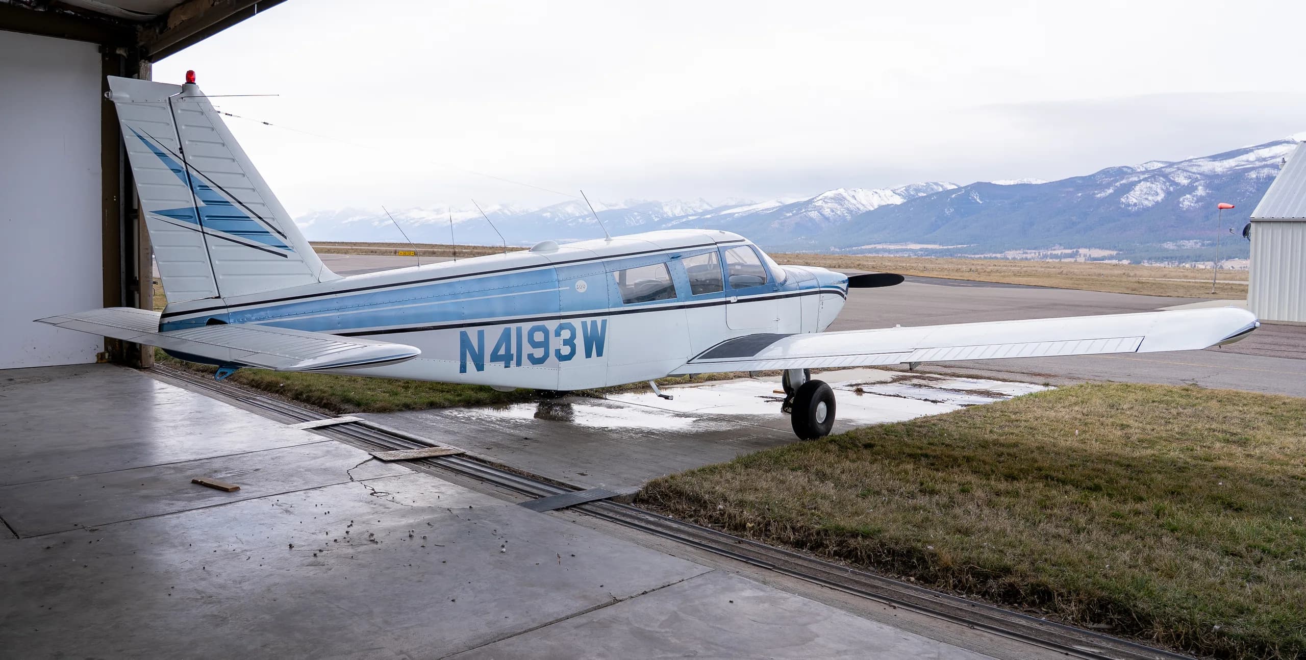 Side view of a white and blue single-engine propeller aircraft N4193W partially in a hangar with snowy mountains in the background.