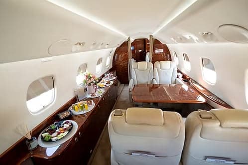 A wide view of the private jet's cabin, showcasing cream leather seating, a large wooden table, and a buffet of food and drinks.