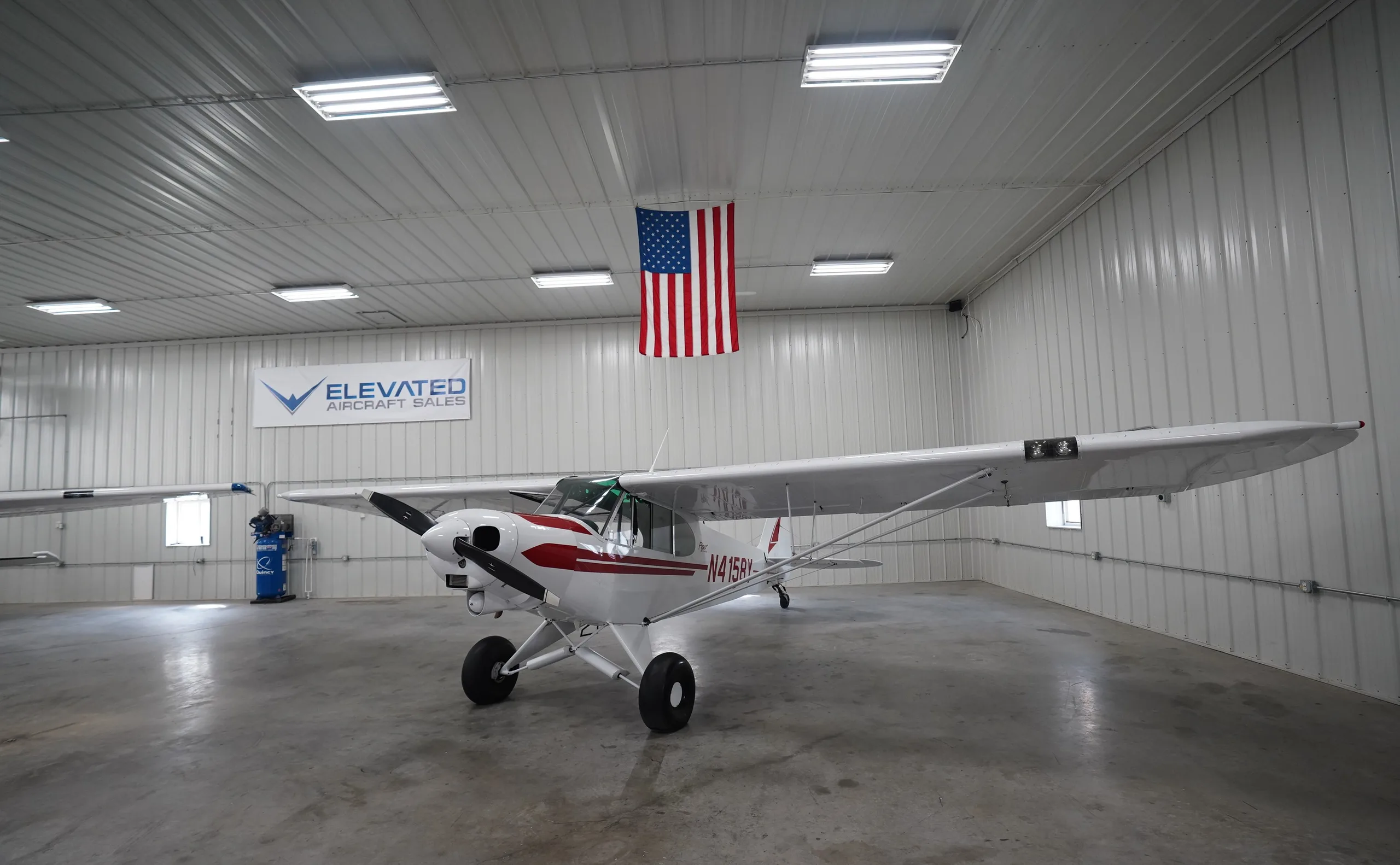 A white Piper Cub aircraft, registration N4158Y, with red stripes is parked in a hangar, with an "Elevated Aircraft Sales" banner and an American flag.