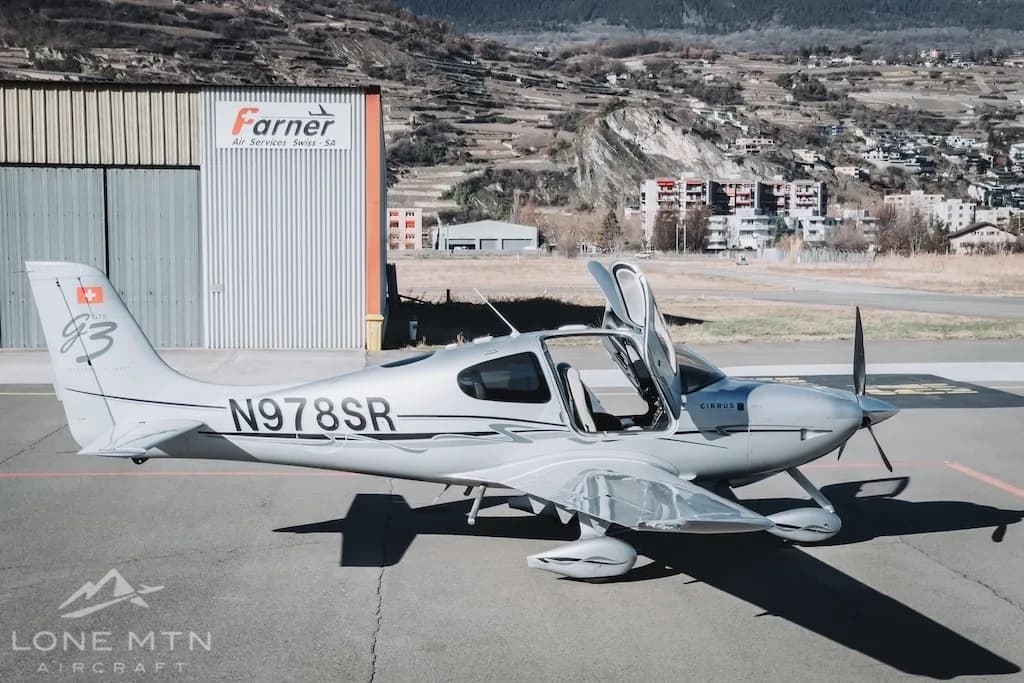 A silver Cirrus SR22 aircraft (N978SR) with its doors open, parked on an airfield next to a hangar.