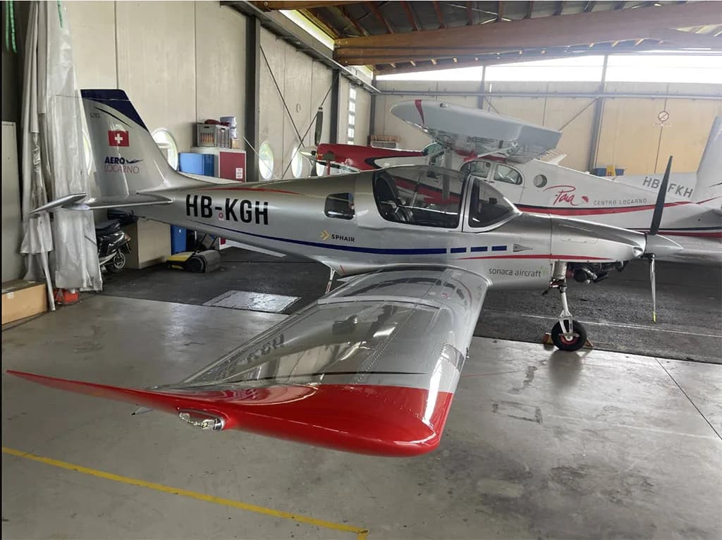 Rear three-quarter view of a silver Sonaca 200 aircraft with red wingtips and blue tail, registration HB-KGH, in a hangar.