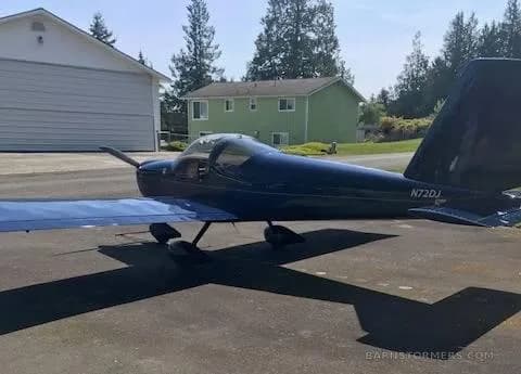 A dark blue single-engine aircraft with tail number N72DJ is parked on a concrete pad with houses and trees in the background.