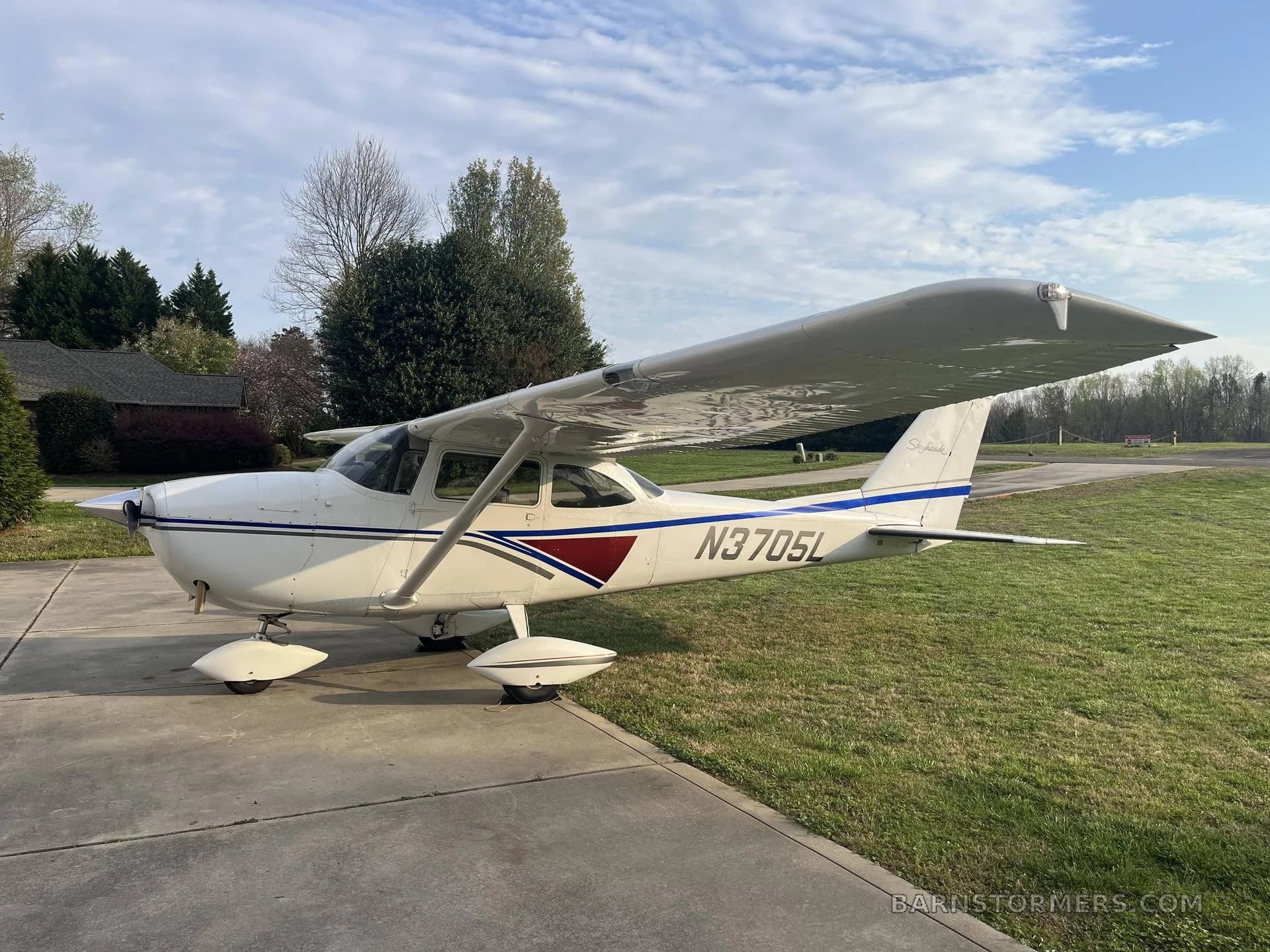 A white Cessna Skyhawk (N3705L) with blue and maroon stripes is parked on a concrete pad next to a grassy area under a partly cloudy sky.