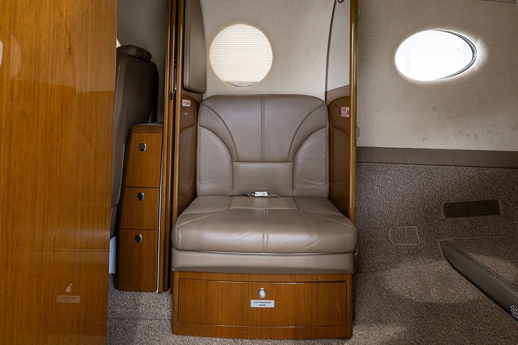 A close-up view of a single beige leather seat and a wooden cabinet in the aircraft's cabin.