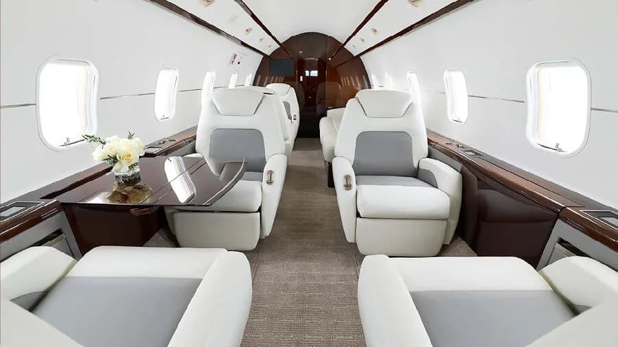 The luxurious interior of a private jet cabin with light gray and white leather seats and dark wood accents.