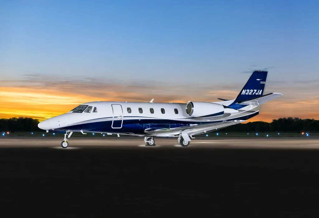 A white and blue Cessna Citation Latitude business jet with registration N327JA on a tarmac at sunset.