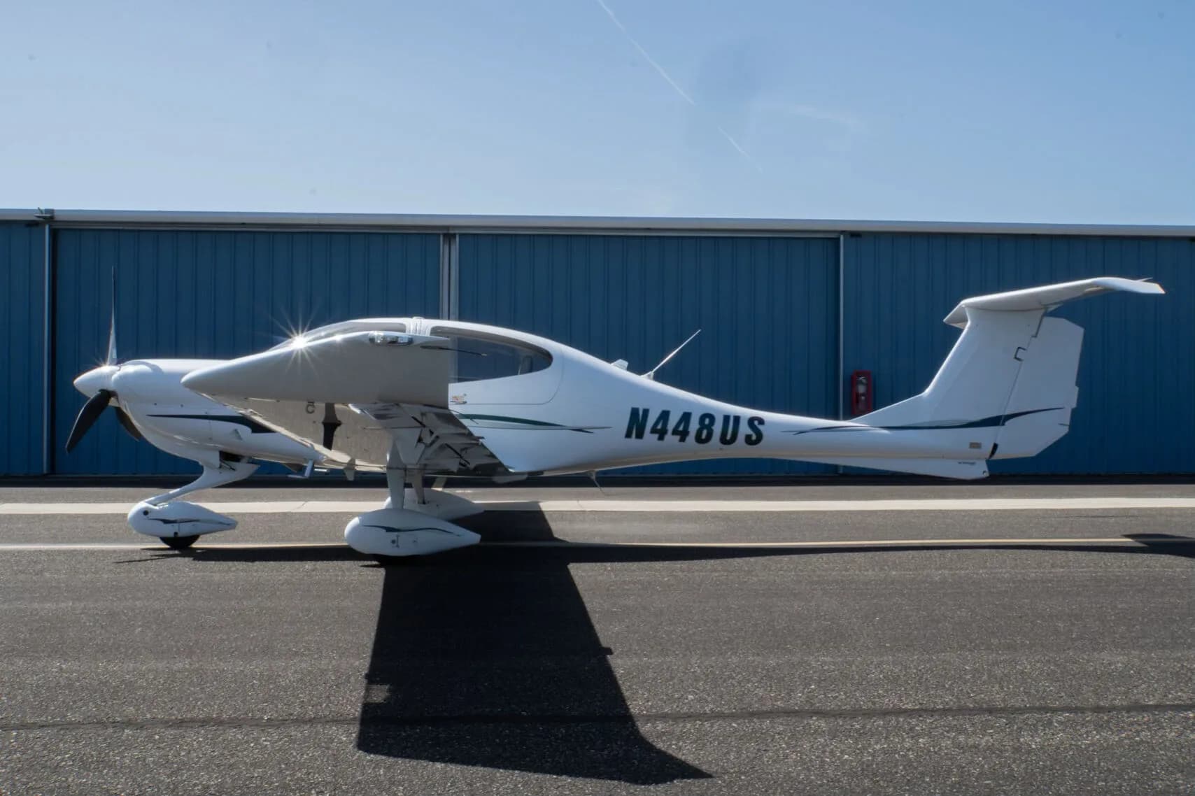 A white Diamond DA40 Star XL aircraft with green and grey stripes, registration N448US, parked on an tarmac in front of a blue hangar.