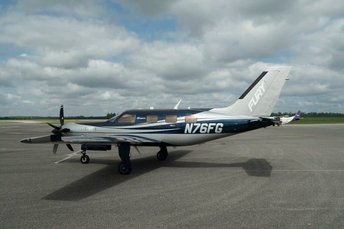 A Piper M700 Fury aircraft with a blue and silver paint scheme, registration N76FG, parked on an airfield under a partly cloudy sky.