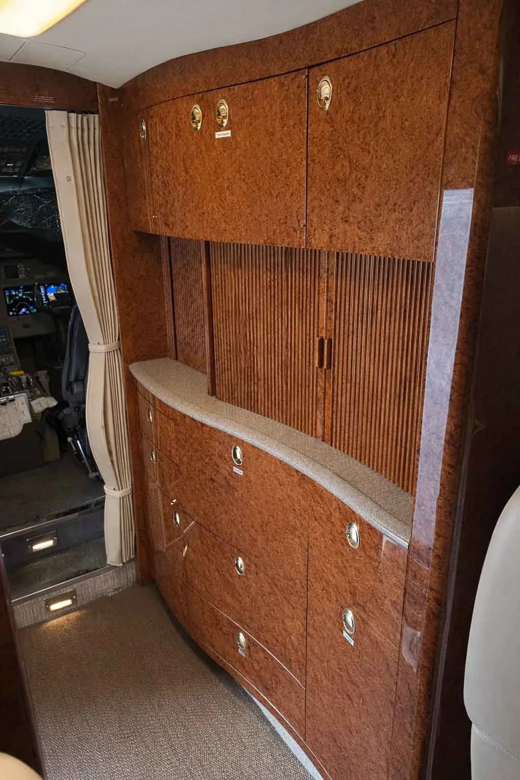 A view of the business jet's forward cabin, showing a wood-paneled galley or storage unit and a glimpse into the cockpit.