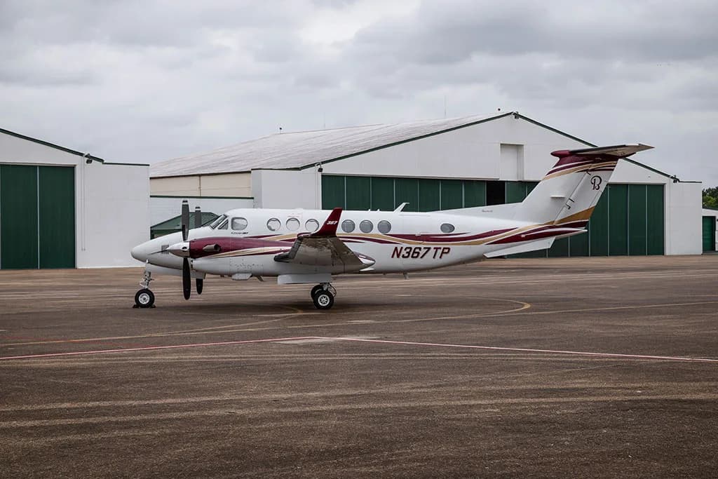 A side view of the N367TP twin-engine turboprop aircraft, white with maroon and gold accents, parked in front of hangars.