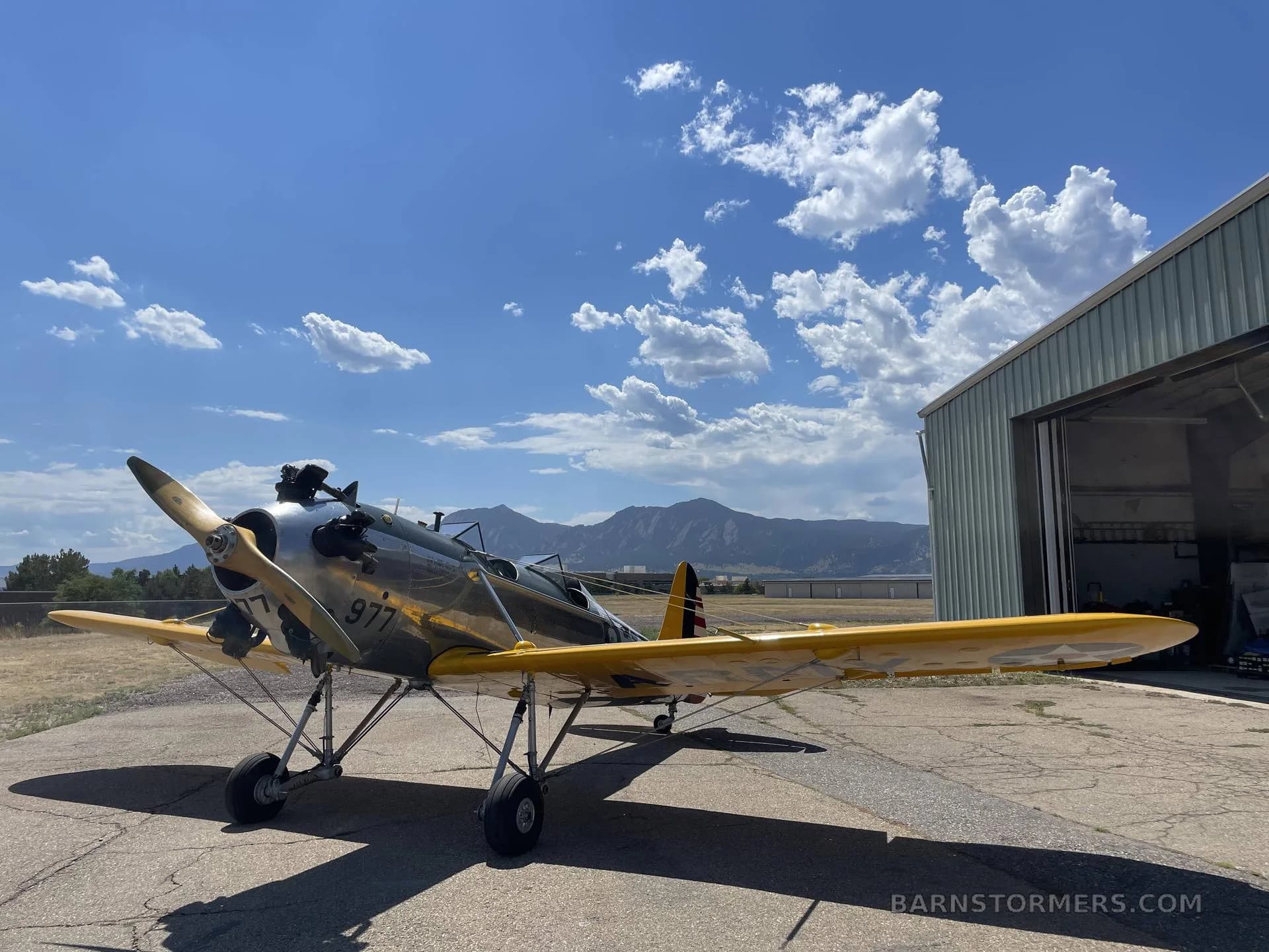 A silver and yellow biplane with '977' on the side, parked on an tarmac next to an open hangar under a blue sky.