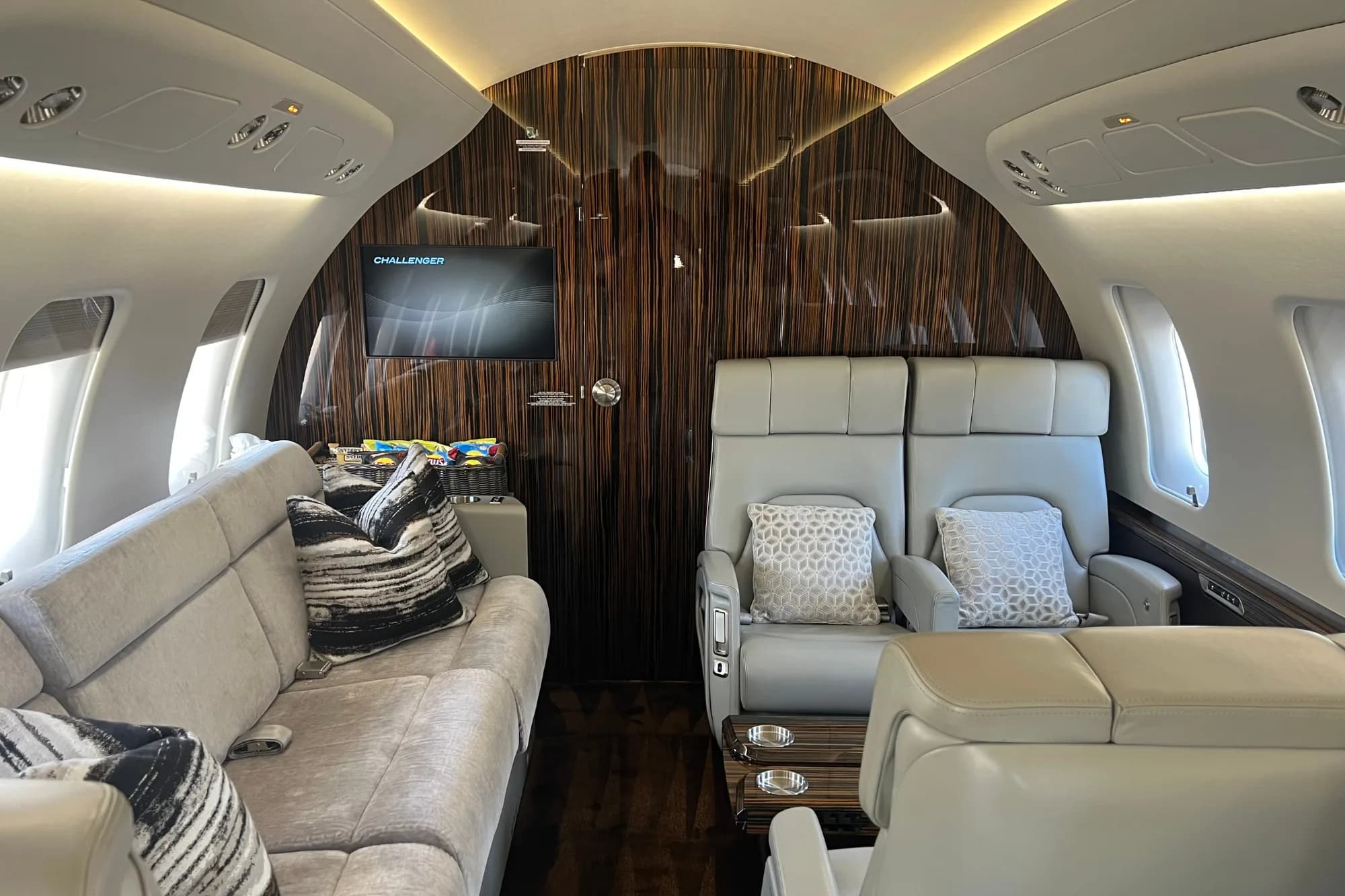 Comfortable aircraft cabin with a grey sofa, two seats, and a screen.