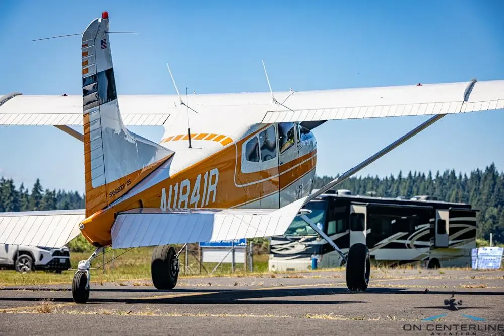 A rear-side view of a white and orange Cessna 185 Skywagon (N1841R) on an asphalt ramp with an RV in the background.