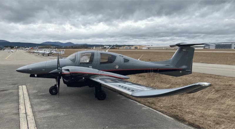 A grey Diamond DA62 twin-engine aircraft with red pinstripes parked on an airfield tarmac with other planes in the background.