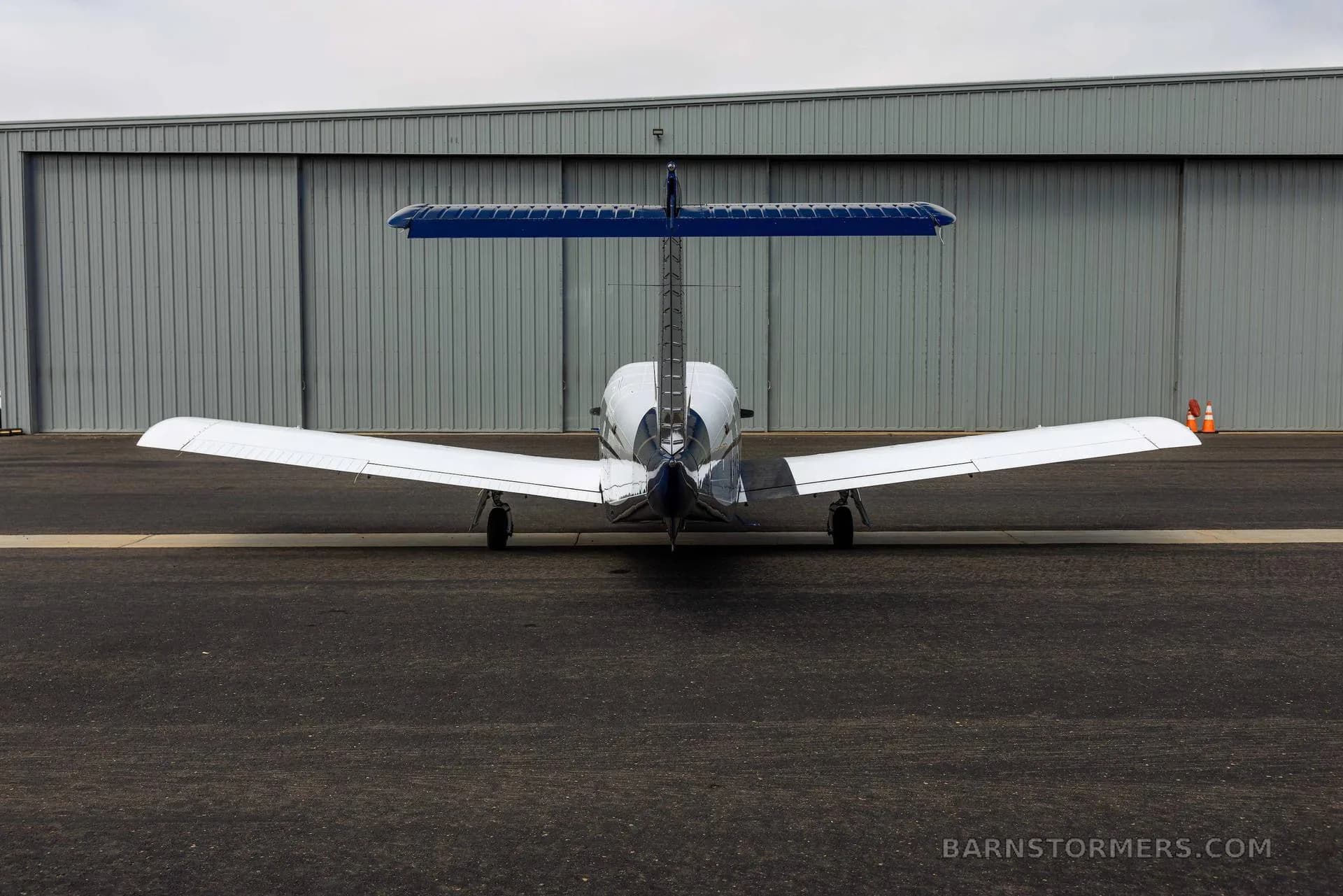A rear view of a white and blue single-engine propeller aircraft with a T-tail, parked on an asphalt tarmac in front of a hangar.