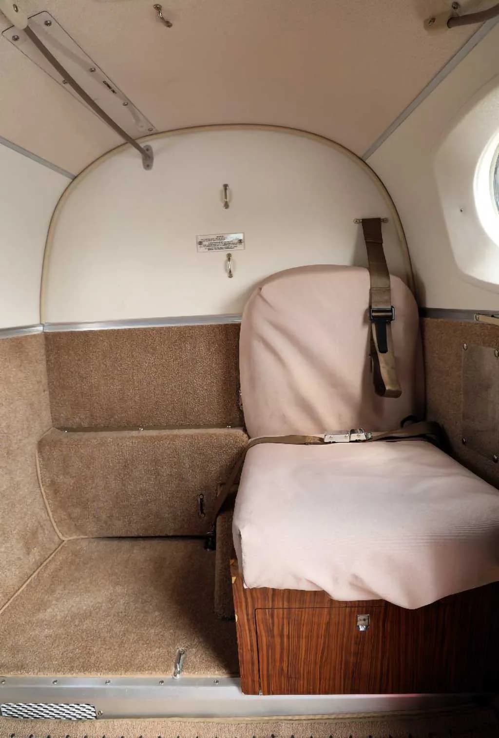 The rear section of an aircraft cabin with a light-colored bench seat and brown carpeted walls.