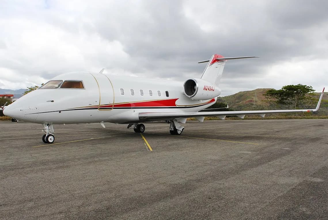 A white Bombardier Challenger business jet with red and gold stripes is parked on an asphalt tarmac under a cloudy sky.