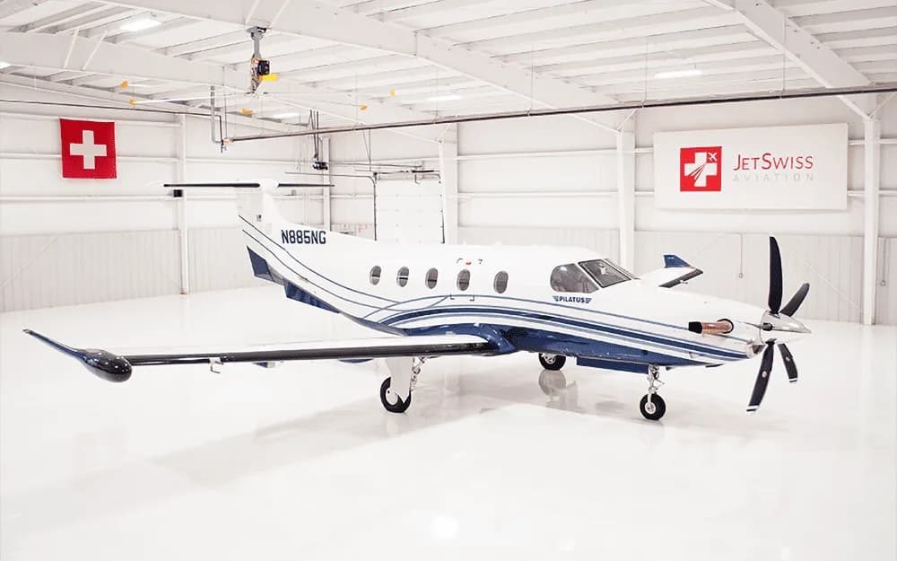 A white and blue Pilatus PC-12 aircraft, tail number N885NG, is parked in a brightly lit hangar, viewed from the front-right.