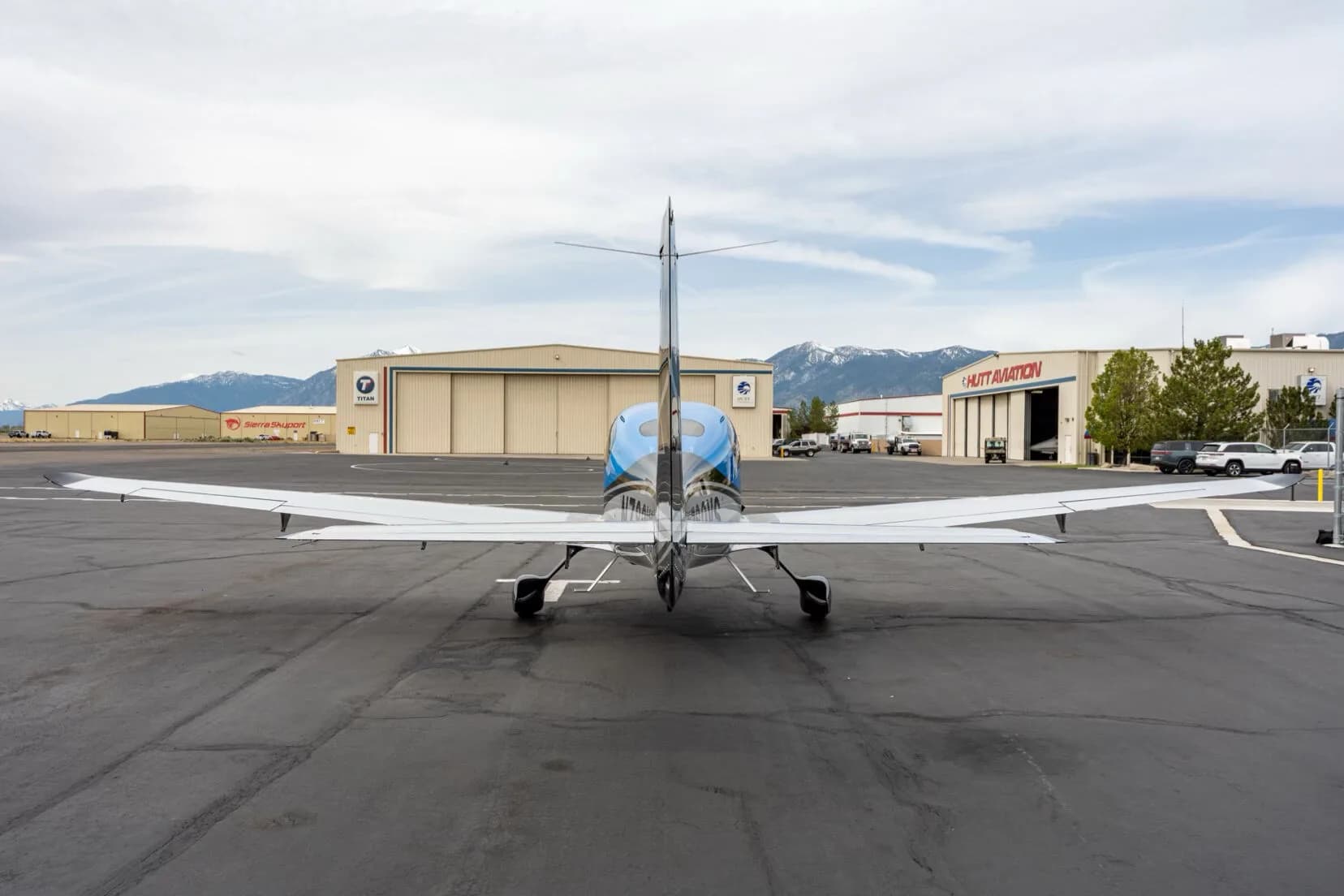 Rear view of a black, blue, and silver single-engine aircraft parked on an airfield with hangars in the background.